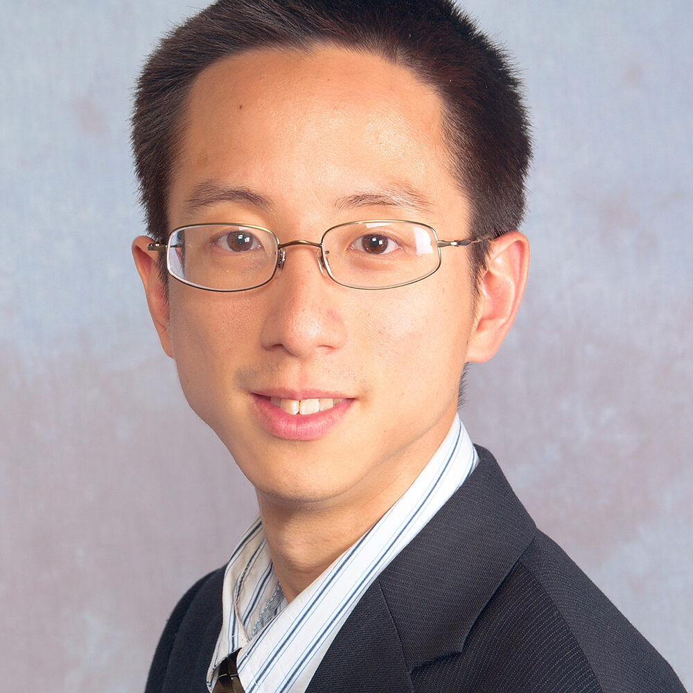 Arthur Chan wearing a dark suit and striped shirt and tie while smiling to camera in front of a pale grey background