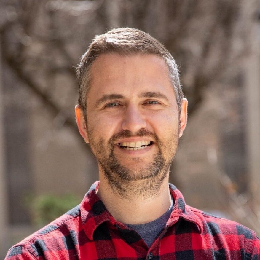 Stephen Johns wearing a red and black checked shirt and smiling to camera outdoors in front of a tree